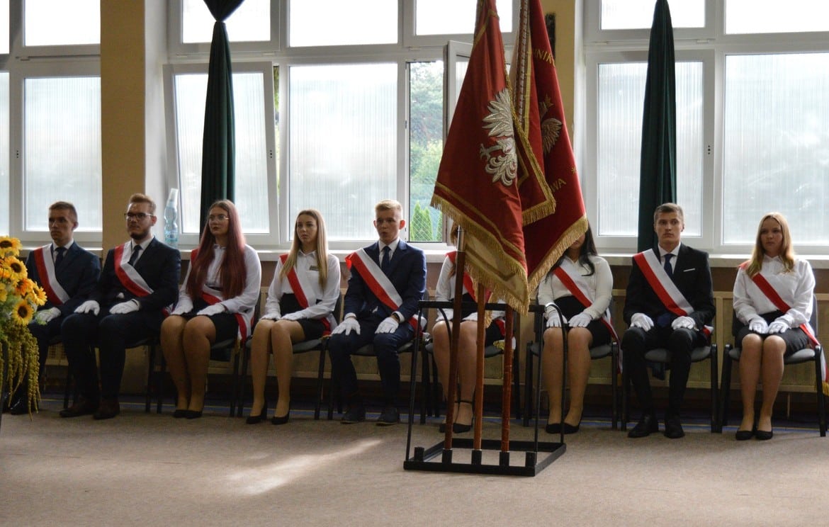 Grupa młodych ludzi w eleganckich strojach z odznakami siedzi w rzędzie, a obok nich stoją sztandary.