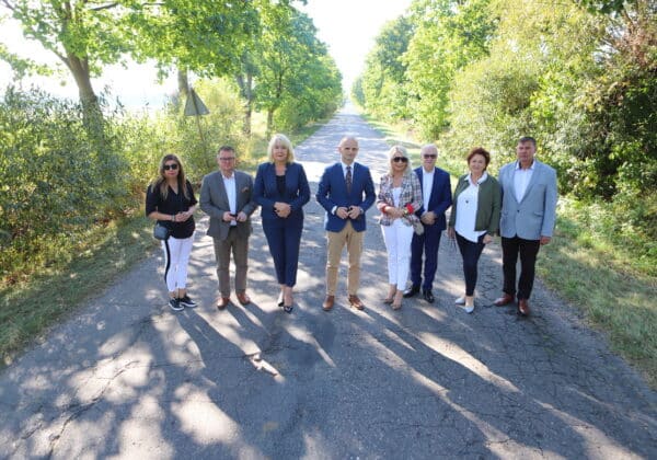 Na zdjęciu widoczna jest grupa ludzi stojących na drodze otoczonej zielenią, w promieniach porannego słońca.