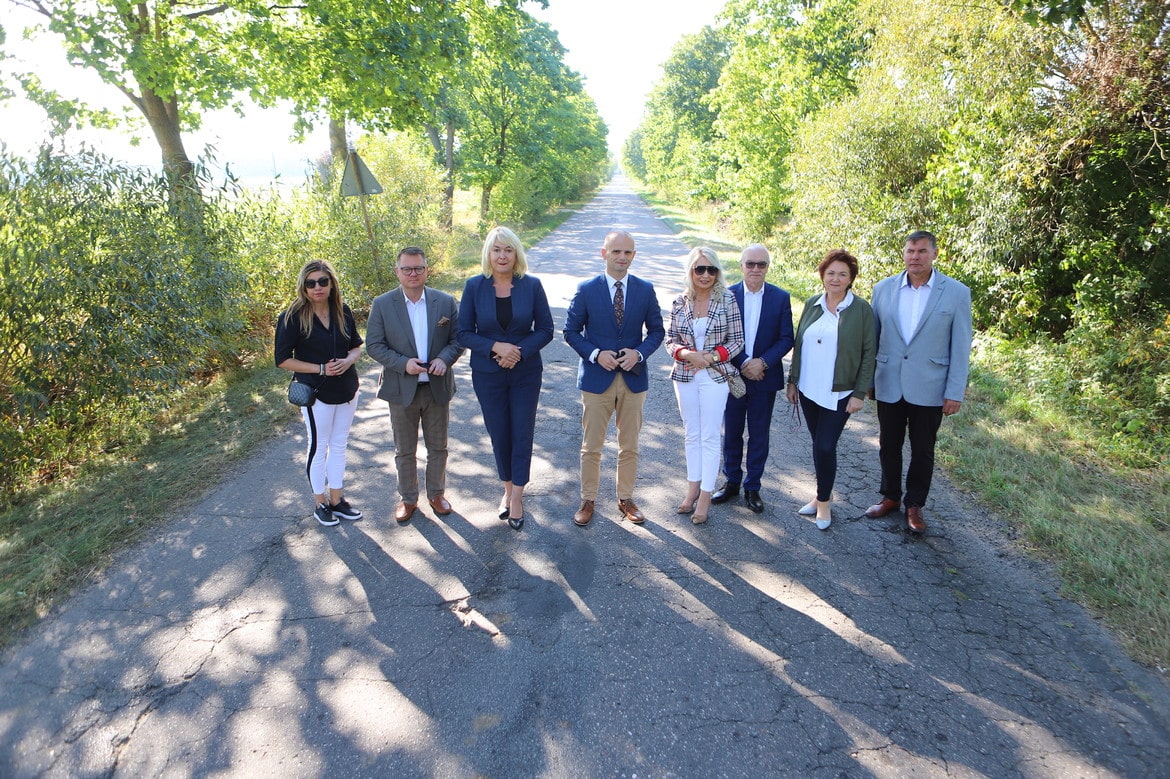 Na zdjęciu widoczna jest grupa ludzi stojących na drodze otoczonej zielenią, w promieniach porannego słońca.