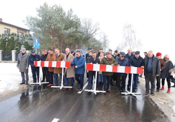 Na zdjęciu widoczna jest grupa osób, w tym lokalnych liderów i przedstawicieli, trzymających barierki drogowe w zimowej scenerii, na tle zaśnieżonych drzew i budynków.