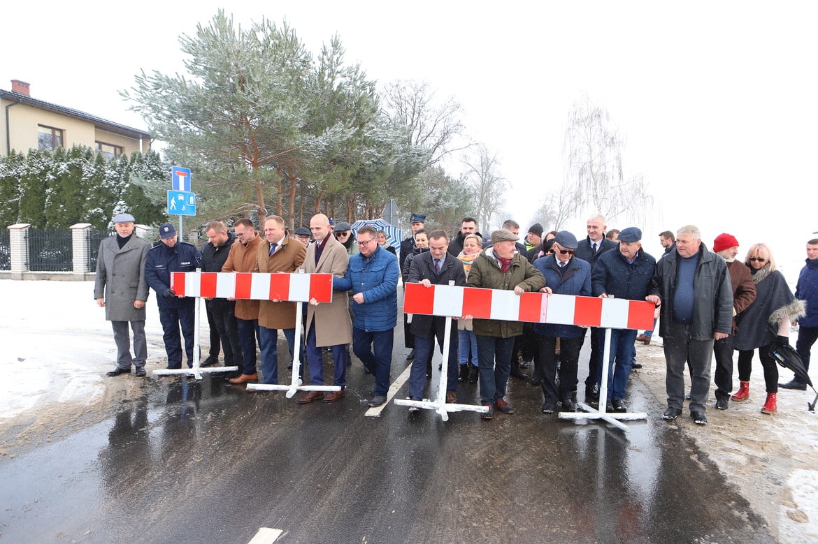 Na zdjęciu widoczna jest grupa osób, w tym lokalnych liderów i przedstawicieli, trzymających barierki drogowe w zimowej scenerii, na tle zaśnieżonych drzew i budynków.