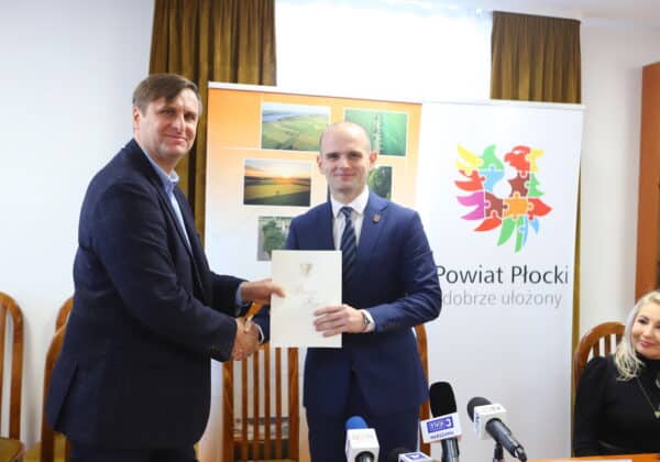Na zdjęciu dwaj mężczyźni uściskiem dłoni dokonują podpisania dokumentu w obecności kobiet i sprzętu medialnego w siedzibie Powiatu Płockiego.