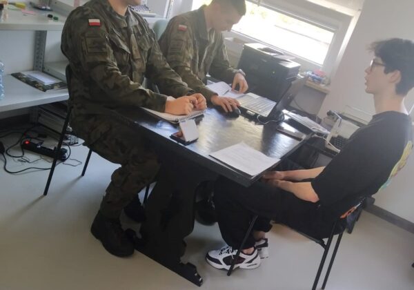 Dwóch żołnierzy w mundurach wojskowych siedzi przy stole, prowadząc rozmowę z młodym mężczyzną.