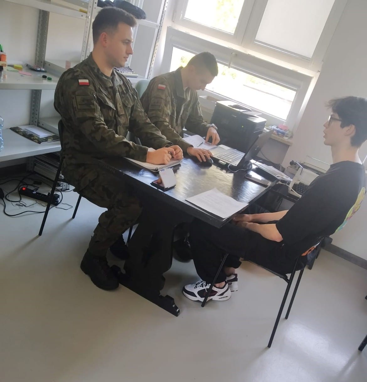 Dwóch żołnierzy w mundurach wojskowych siedzi przy stole, prowadząc rozmowę z młodym mężczyzną.