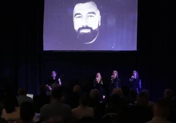 Na zdjęciu widać występ czterech artystek śpiewających na scenie, z dużym ekranem w tle, na którym wyświetlona jest twarz mężczyzny.