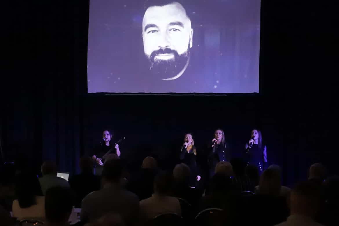 Na zdjęciu widać występ czterech artystek śpiewających na scenie, z dużym ekranem w tle, na którym wyświetlona jest twarz mężczyzny.