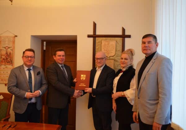 Na zdjęciu przedstawiono cztery osoby stojące w pomieszczeniu, które trzymają dokument oraz uściskiem dłoni potwierdzają współpracę.