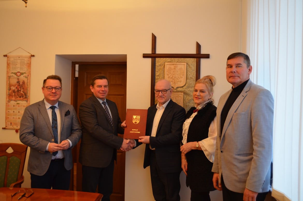 Na zdjęciu przedstawiono cztery osoby stojące w pomieszczeniu, które trzymają dokument oraz uściskiem dłoni potwierdzają współpracę.