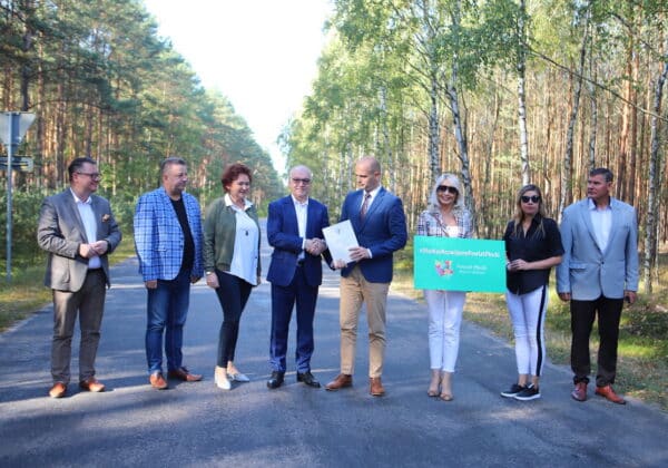 Na zdjęciu w otoczeniu drzew grupka ludzi stoi na drodze, podpisując dokumenty oraz trzymając transparent z hasłem promującym rozwój powiatu płockiego.