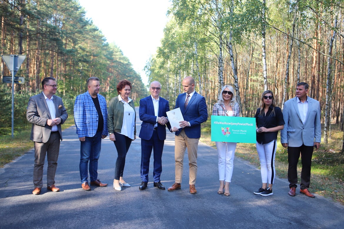 Na zdjęciu w otoczeniu drzew grupka ludzi stoi na drodze, podpisując dokumenty oraz trzymając transparent z hasłem promującym rozwój powiatu płockiego.