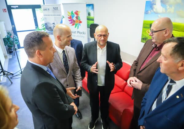 Na zdjęciu widoczna jest grupa mężczyzn dyskutujących w jasnym pomieszczeniu, w otoczeniu bannerów promujących powiat płocki.