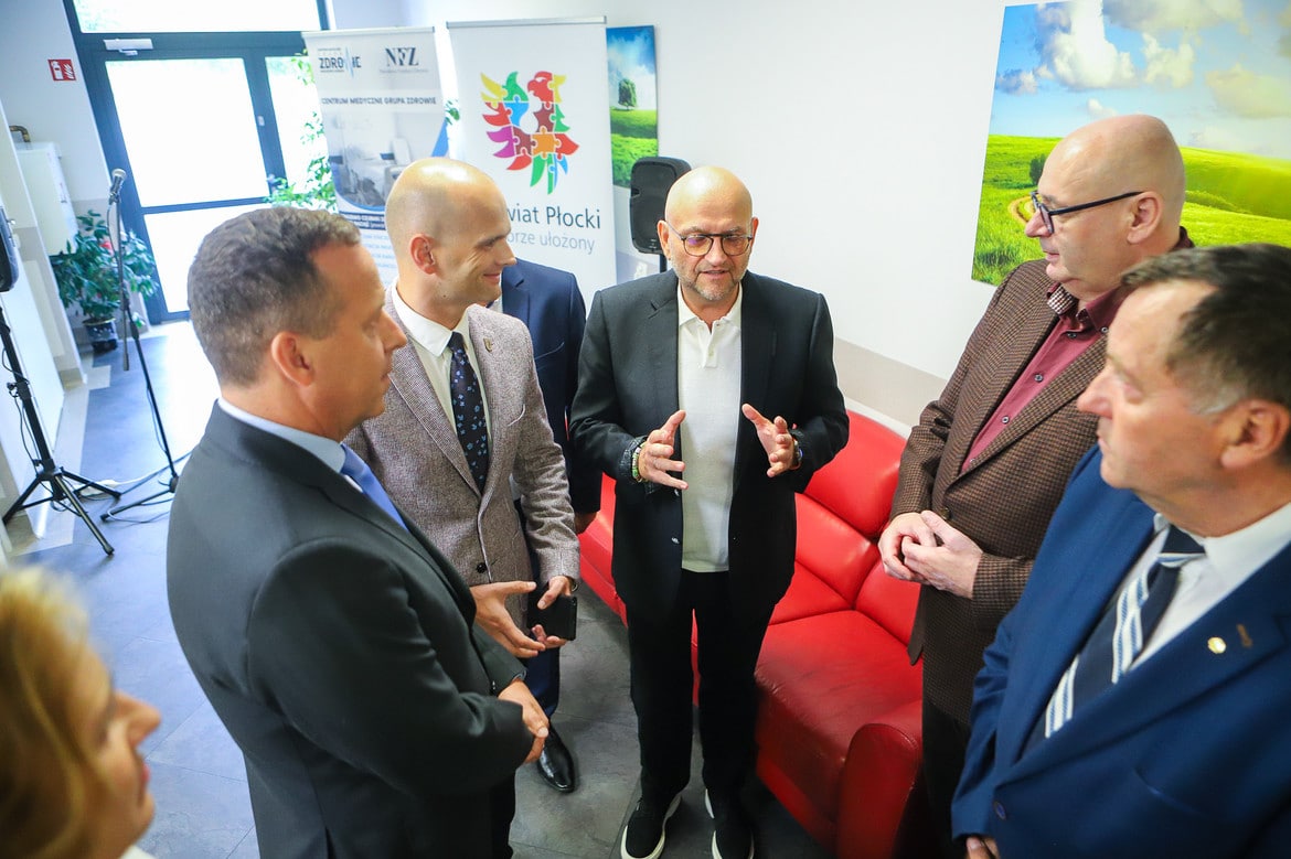 Na zdjęciu widoczna jest grupa mężczyzn dyskutujących w jasnym pomieszczeniu, w otoczeniu bannerów promujących powiat płocki.