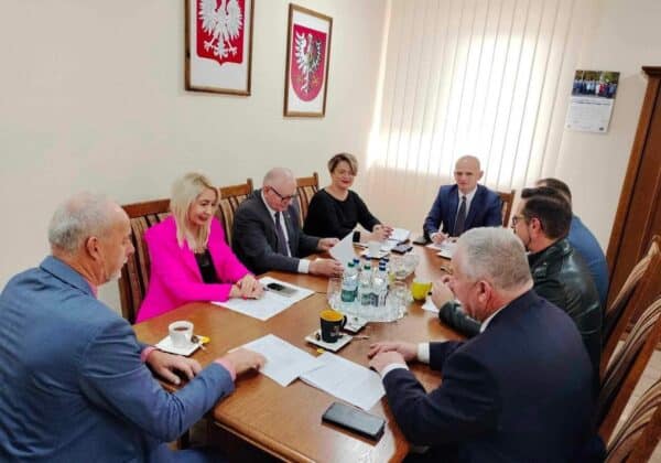 Spotkanie grupy osób w formalnym otoczeniu, siedzących przy stole z dokumentami, napotykających na obrazy godła Polski na ścianie.