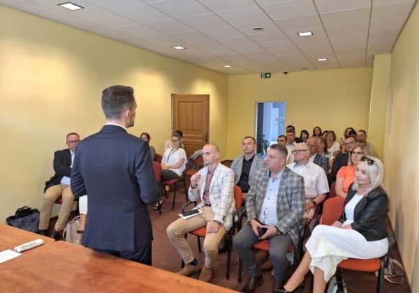 Zdjęcie przedstawia grupę osób siedzących w sali konferencyjnej, słuchających prezentacji wygłaszanej przez mężczyznę w garniturze.