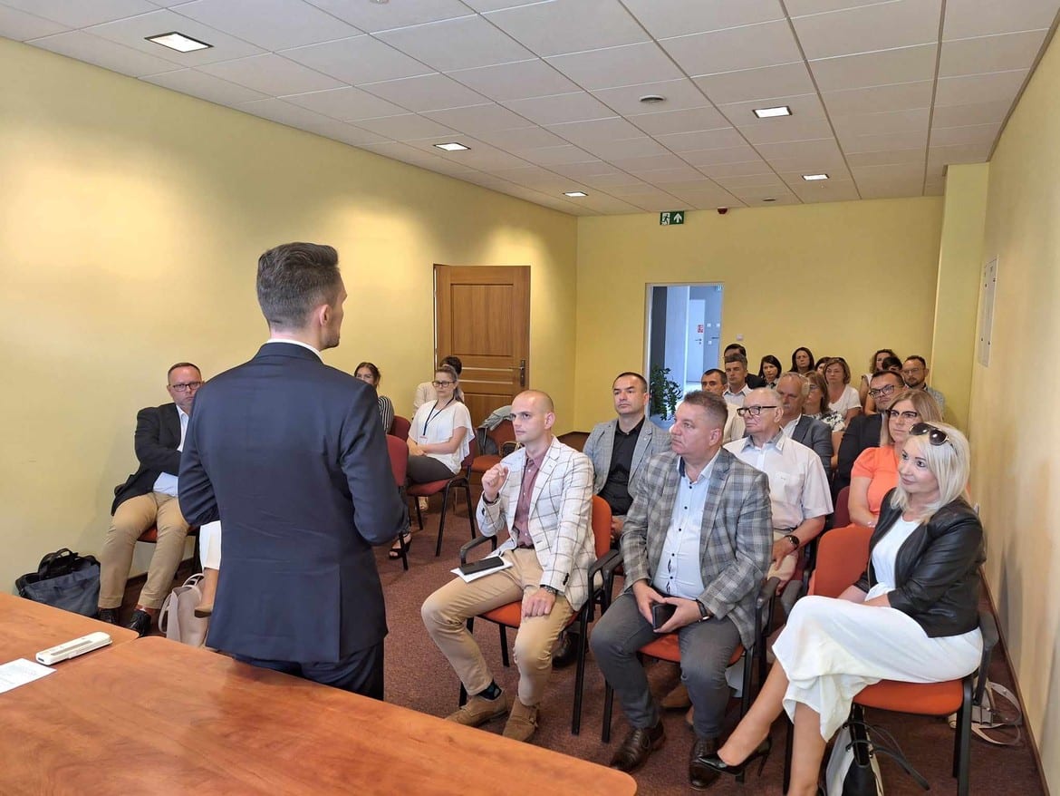 Zdjęcie przedstawia grupę osób siedzących w sali konferencyjnej, słuchających prezentacji wygłaszanej przez mężczyznę w garniturze.