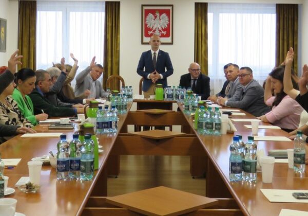 Na zdjęciu przedstawiono zebranie, na którym uczestnicy podnoszą ręce w celu głosowania, a przewodniczący stoi na czołowej pozycji przy stole.