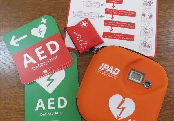 Na zdjęciu znajdują się materiały związane z defibrylatorem AED, w tym instrukcje oraz sam defibrylator w kolorze pomarańczowym.