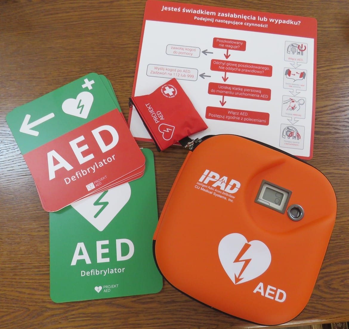 Na zdjęciu znajdują się materiały związane z defibrylatorem AED, w tym instrukcje oraz sam defibrylator w kolorze pomarańczowym.