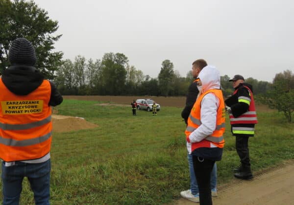 Na zdjęciu grupa osób w kamizelkach odblaskowych obserwuje akcję ratunkową na otwartym terenie.