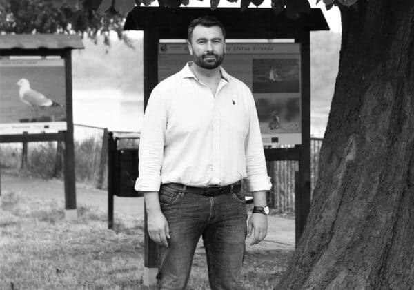 Mężczyzna w białej koszuli i dżinsach stoi na trawie przed tablicami informacyjnymi w parku.