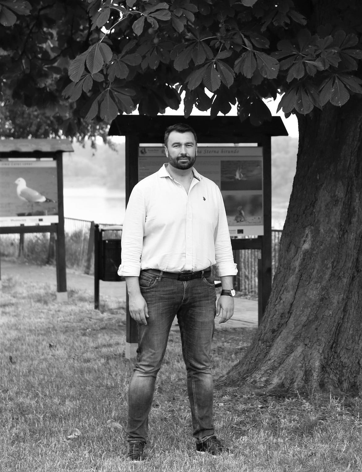 Mężczyzna w białej koszuli i dżinsach stoi na trawie przed tablicami informacyjnymi w parku.