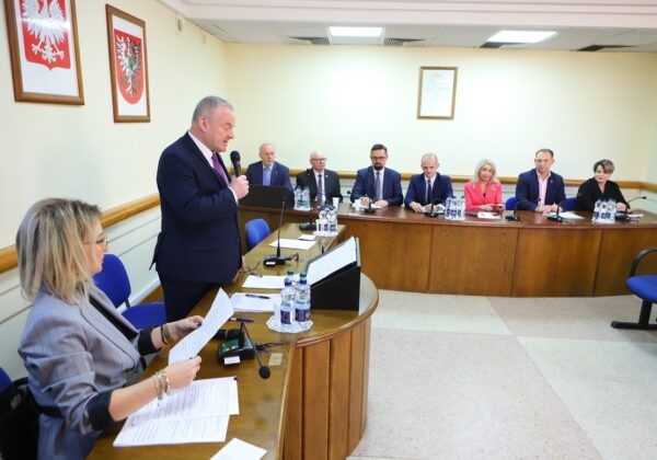 Na zdjęciu przedstawiono zebranie w sali konferencyjnej, podczas którego mężczyzna przemawia do siedzących przy stole uczestników spotkania.