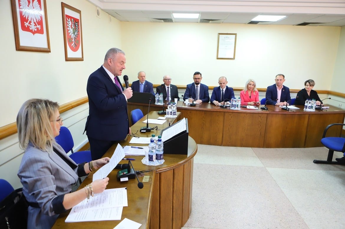 Na zdjęciu przedstawiono zebranie w sali konferencyjnej, podczas którego mężczyzna przemawia do siedzących przy stole uczestników spotkania.