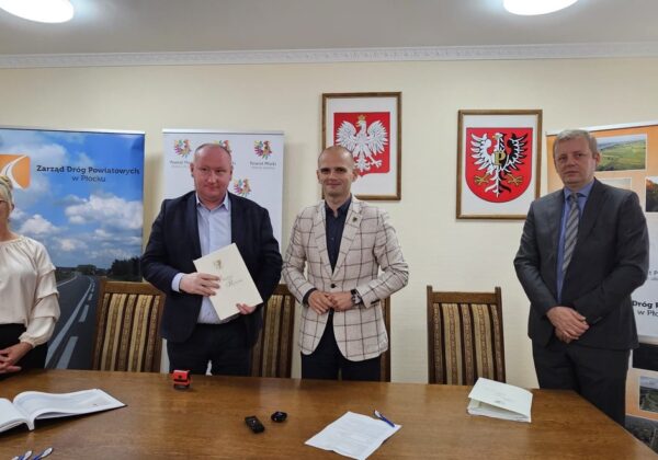 Na zdjęciu przedstawiono czterech mężczyzn i kobietę stojących przy stole, trzymających dokumenty, w tle widoczne banery promujące Powiat Płocki oraz Zarząd Dróg Powiatowych w Płocku.