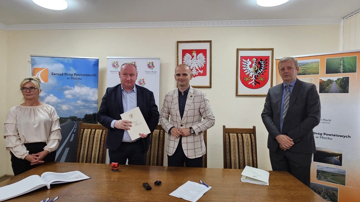 Na zdjęciu przedstawiono czterech mężczyzn i kobietę stojących przy stole, trzymających dokumenty, w tle widoczne banery promujące Powiat Płocki oraz Zarząd Dróg Powiatowych w Płocku.