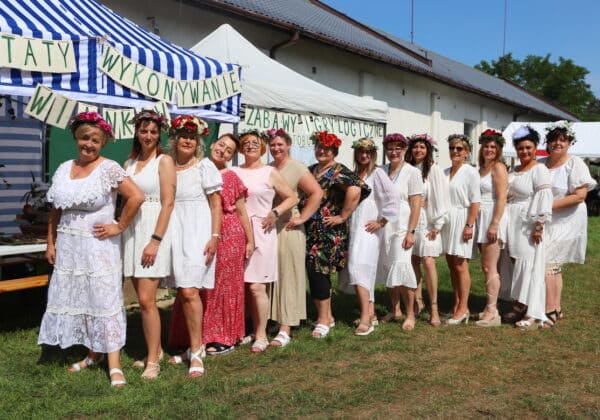 Na zdjęciu znajduje się grupa kobiet ubranych w białe sukienki, w kwiatach we włosach, pozująca przed namiotem z napisami „Warsztaty wykonywanie winka” i „Zabawy i gry logiczne”.