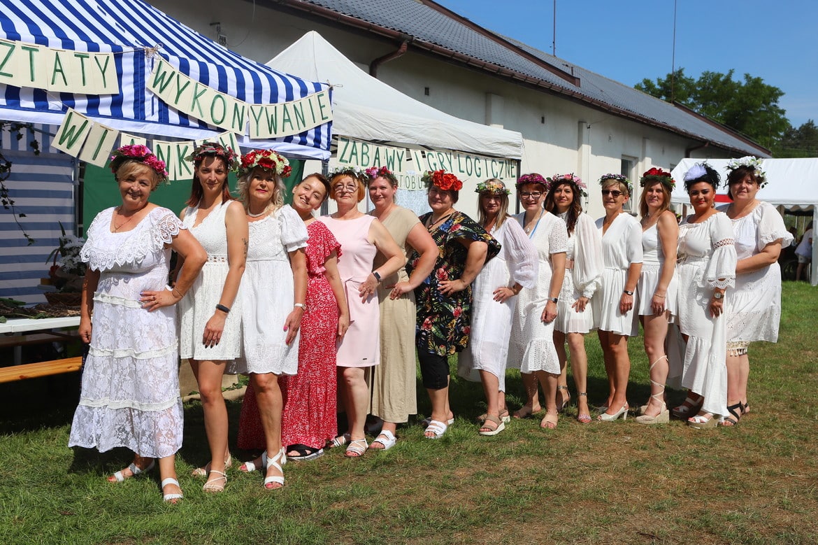 Na zdjęciu znajduje się grupa kobiet ubranych w białe sukienki, w kwiatach we włosach, pozująca przed namiotem z napisami „Warsztaty wykonywanie winka” i „Zabawy i gry logiczne”.