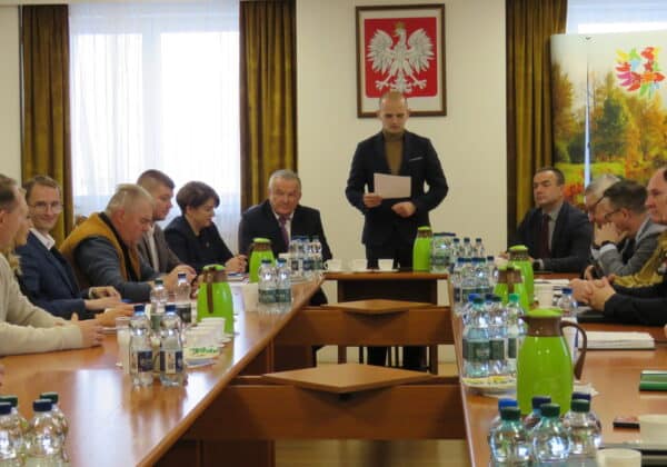 Na zdjęciu przedstawiono spotkanie grupy osób w sali konferencyjnej, w której omawiane są ważne sprawy, z widocznym prowadzącym i osobami siedzącymi przy stole.