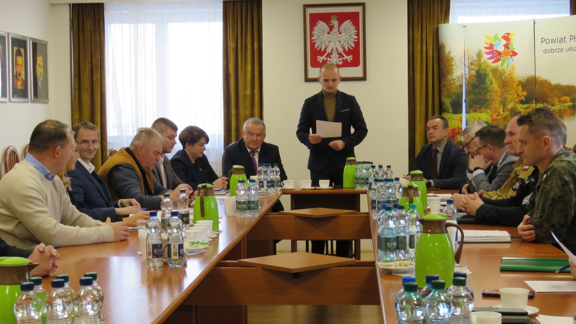 Na zdjęciu przedstawiono spotkanie grupy osób w sali konferencyjnej, w której omawiane są ważne sprawy, z widocznym prowadzącym i osobami siedzącymi przy stole.