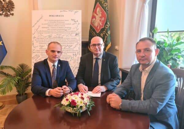 Na zdjęciu trzej mężczyźni siedzą przy stole w eleganckim pomieszczeniu, z tłem zawierającym dokument oraz flagi, w ramach podpisania ważnej deklaracji.