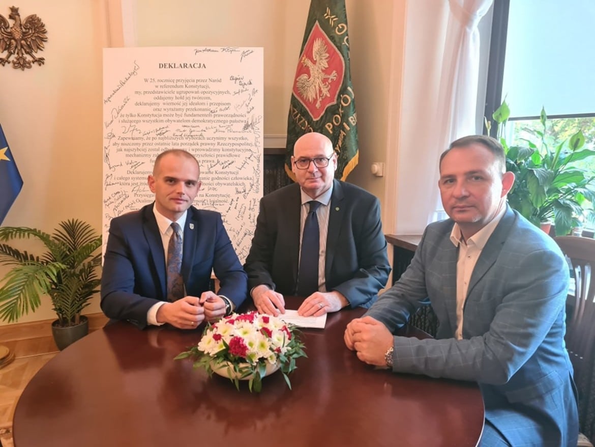 Na zdjęciu trzej mężczyźni siedzą przy stole w eleganckim pomieszczeniu, z tłem zawierającym dokument oraz flagi, w ramach podpisania ważnej deklaracji.