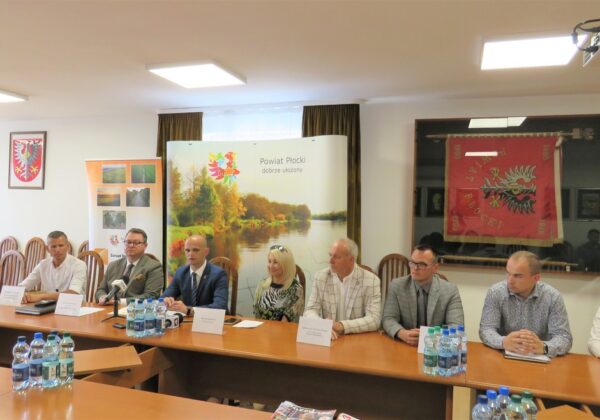 Na zdjęciu przedstawiono grupę osób siedzących przy stole konferencyjnym w sali obrad, z banerem promującym powiat płocki w tle.