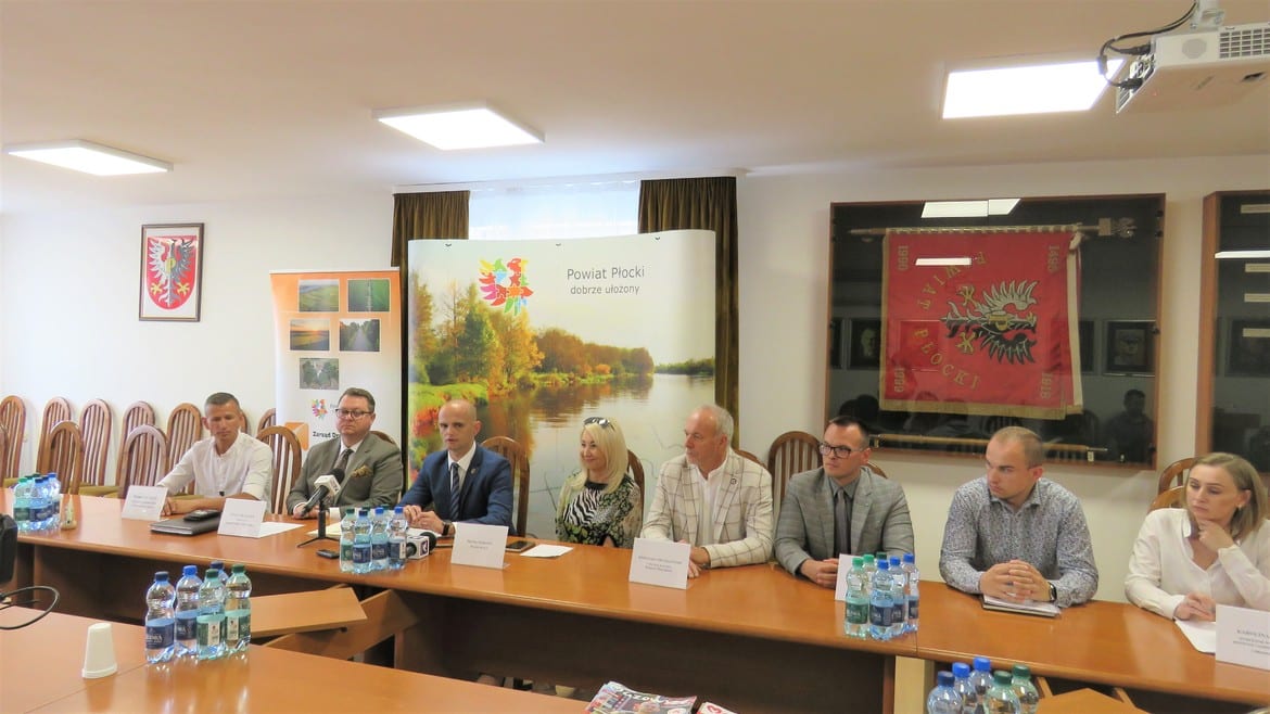 Na zdjęciu przedstawiono grupę osób siedzących przy stole konferencyjnym w sali obrad, z banerem promującym powiat płocki w tle.