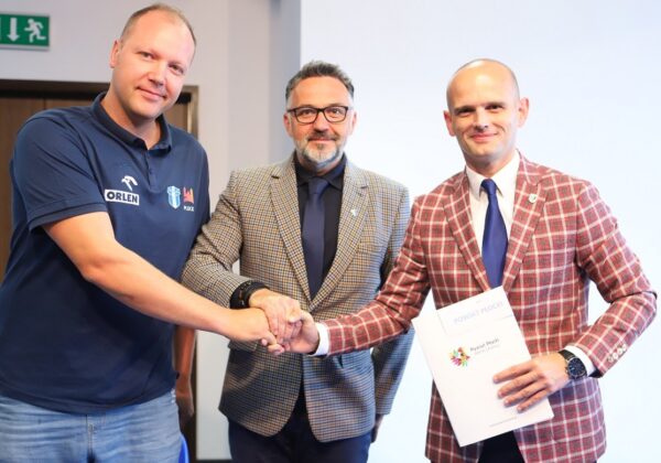 Na zdjęciu trzej mężczyźni uścisną ręce, podczas uroczystości wręczenia dokumentu w sali konferencyjnej.