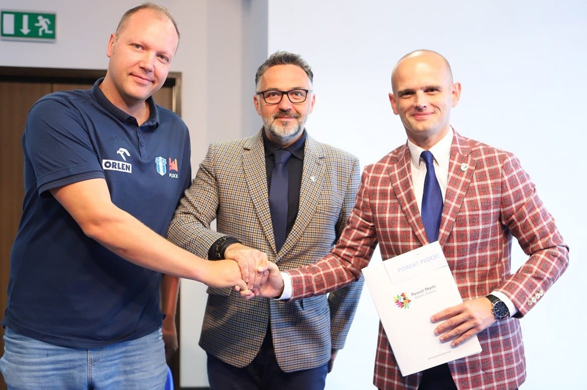 Na zdjęciu trzej mężczyźni uścisną ręce, podczas uroczystości wręczenia dokumentu w sali konferencyjnej.