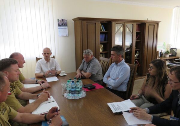 Na zdjęciu grupa osób, w tym mężczyźni w mundurach, prowadzi dyskusję przy stole w jasnym pomieszczeniu biurowym.