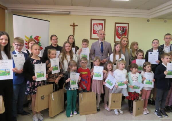 Na zdjęciu grupa dzieci z dyplomami stoi w sali, uśmiechając się do aparatu, obok dorosłych, w tle widoczne flagi i insygnia.