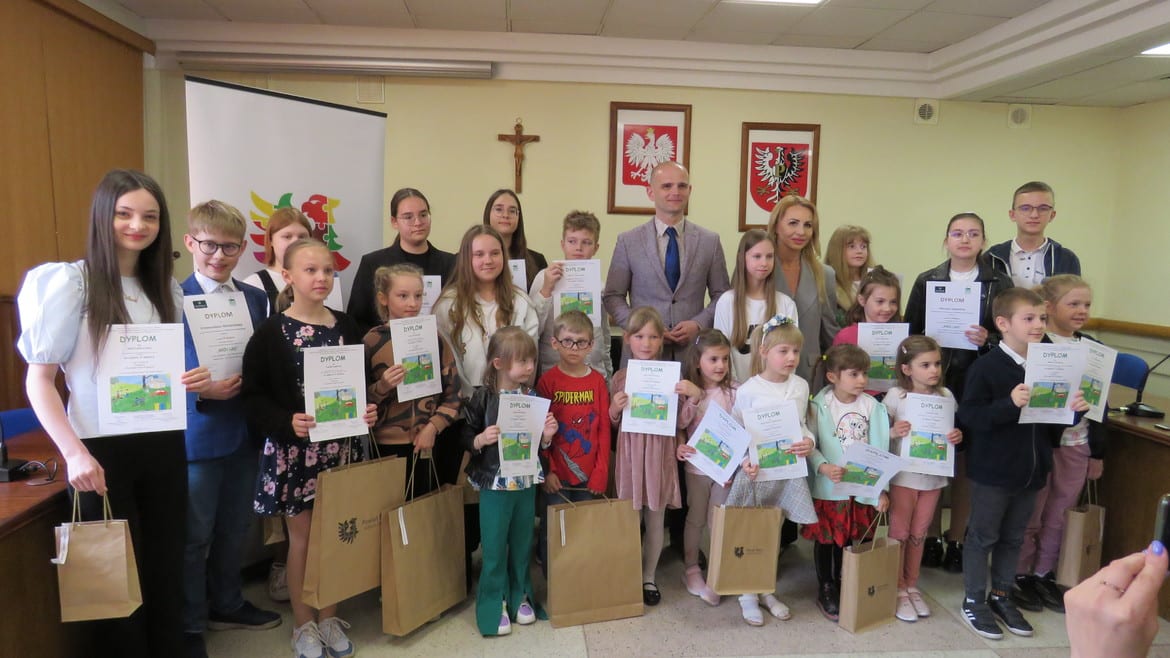 Na zdjęciu grupa dzieci z dyplomami stoi w sali, uśmiechając się do aparatu, obok dorosłych, w tle widoczne flagi i insygnia.
