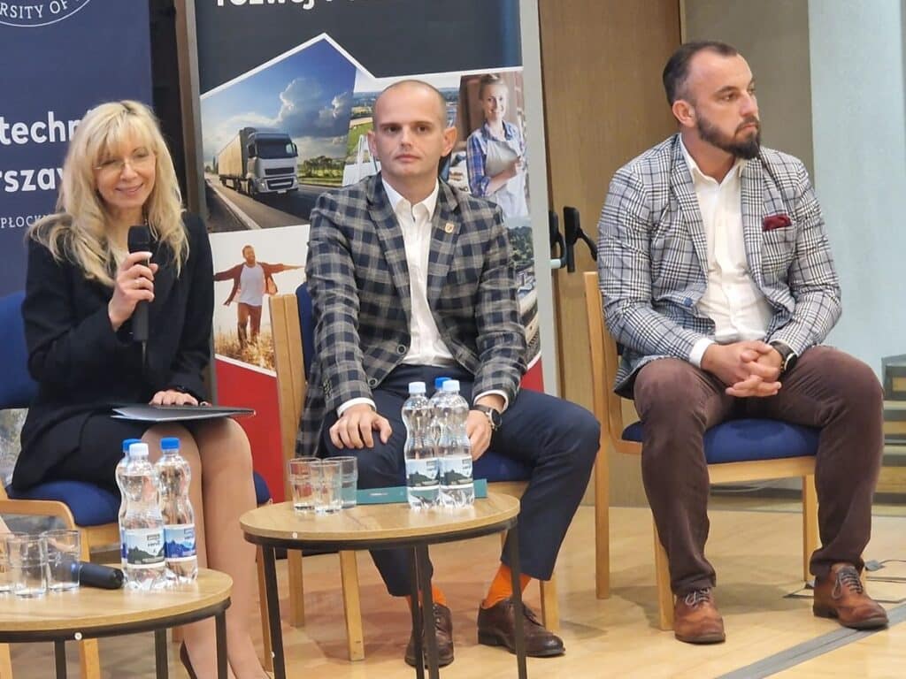 Na zdjęciu przedstawiono trzech uczestników debaty, z których jedna osoba trzyma mikrofon, a dwie pozostałe siedzą obok, w eleganckich strojach.