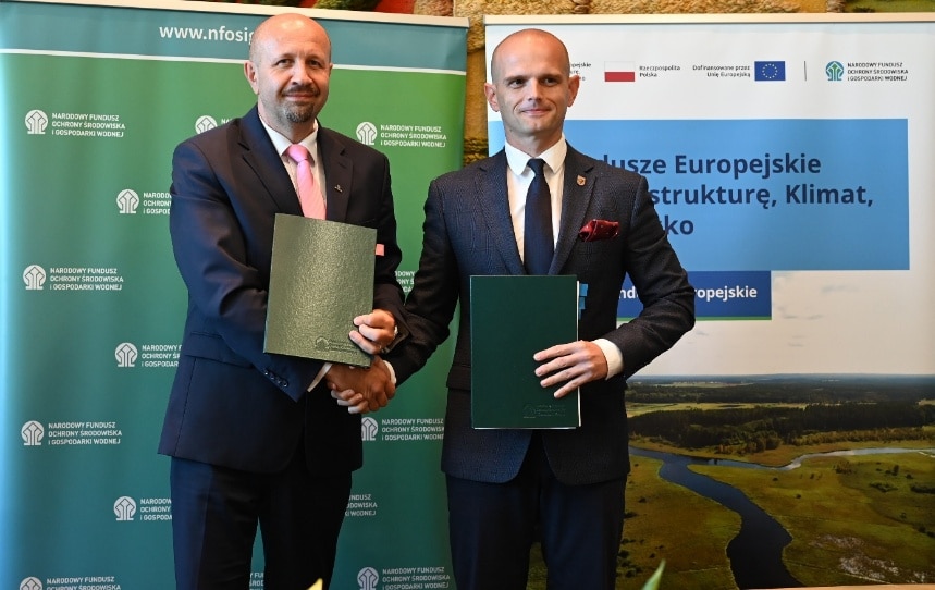 Dwaj mężczyźni, ubrani w formalne stroje, pozują do zdjęcia, trzymając dokumenty w rękach, na tle reklamującym programy ochrony środowiska oraz poprawy systemu przyrodniczego w powiecie płockim.