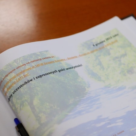 Dokument na stole, dotyczący realizacji inwestycji w gminie, z widokiem na alejkę otoczoną drzewami.