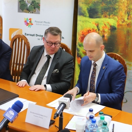 Na zdjęciu przedstawiono dwóch mężczyzn podczas spotkania, siedzących przy stole, przy którym znajdują się dokumenty i mikrofony.