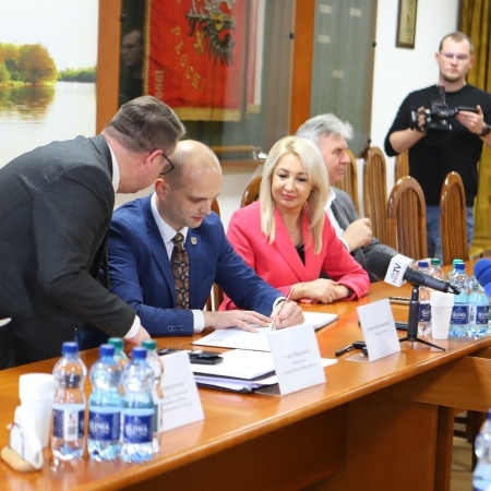 Na zdjęciu przedstawiono spotkanie, podczas którego podpisywana jest umowa, a uczestnicy siedzą przy stole w sali konferencyjnej.