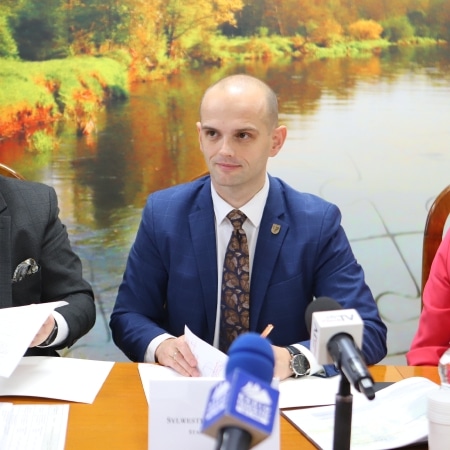 Na zdjęciu przedstawiono mężczyznę w garniturze siedzącego przy stole na tle malowniczej scenerii z wodą i zielenią, w otoczeniu innych osób trzymających dokumenty i mikrofony.