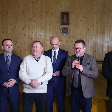 Na zdjęciu widać grupę mężczyzn w formalnych strojach, którzy występują na scenie podczas uroczystości, trzymając mikrofon.