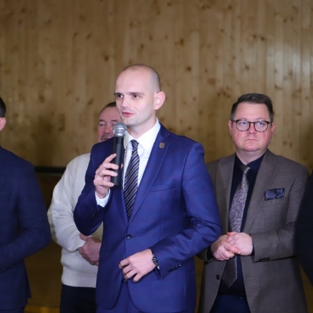 Na zdjęciu widoczny jest mężczyzna w garniturze, trzymający mikrofon i przemawiający do grupy osób w tle na tle drewnianej ściany.